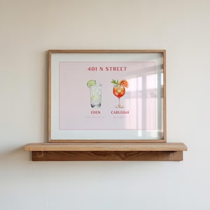 May include: A framed print displaying two cocktail illustrations, "Eden" and "Carleigh", with garnishes, against a pink backdrop. The print includes the text "401 N STREET". The frame is light brown, and the print is on a wooden shelf.