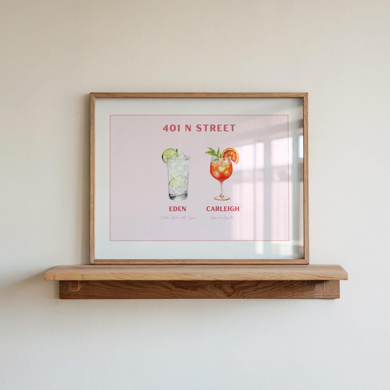 May include: A framed print displaying two cocktail illustrations, "Eden" and "Carleigh", with garnishes, against a pink backdrop. The print includes the text "401 N STREET". The frame is light brown, and the print is on a wooden shelf.