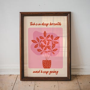 May include: A framed wall art print featuring a potted plant illustration in red on a pink background. The text "Take a deep breath" is printed above the plant, and "and keep going" is printed below, both in a red, handwritten-style font.