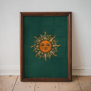 May include: Framed art print with a vintage illustration of a smiling sun in orange and yellow. The sun is set against a dark green background and framed in a dark brown wooden frame. The artwork is displayed on a wooden floor.
