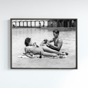May include: A black and white photo of a couple relaxing by a pool. The woman is lying on a towel in a swimsuit, while the man is sitting on the towel with his arm around her. They are both smiling and looking at each other. There is a bottle of beer on the towel between them.
