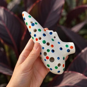 Puede incluir: Una ocarina de cerámica blanca con un acabado de esmalte craquelado y un patrón de pequeños puntos espaciados uniformemente en tonos vibrantes de azul, verde y naranja. El instrumento musical tiene varios orificios en su superficie curva.