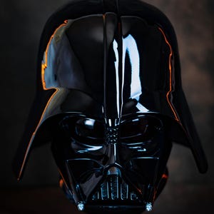 Puede incluir: Un casco negro brillante de Darth Vader, una pieza destacada de Star Wars. La superficie lisa y reflectante del casco captura la luz, resaltando su forma icónica y sus intrincados detalles. El color oscuro y el diseño evocan la presencia amenazante del personaje.