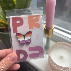 May include: A butterfly pin with a lesbian pride flag design is displayed in a clear plastic bag. The pin features a red, orange, white, and purple colour scheme. The bag is held in front of a window with a pink candle and a small candle in a glass jar.