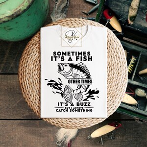 Könnte beinhalten: Weißes T-Shirt mit einem schwarz-weißen Grafikdesign eines Fisches, der aus dem Wasser springt. Der Text lautet "Sometimes it's a fish, other times it's a buzz, but I always catch something."