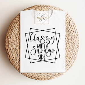 May include: White t-shirt with the text "Classy with a Savage Side" in a black geometric design. The shirt is folded on a woven, circular surface. The brand logo is in the upper left corner.