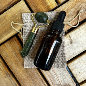 May include: A jade facial roller with gold-colored accents and a brown glass bottle with a black dropper are displayed on a small burlap bag. The items are arranged on a wooden surface, suggesting a skincare or beauty product theme.