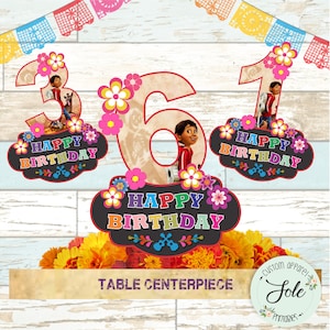 May include: A table centrepiece featuring a large number "6" with a picture of Miguel from the movie Coco, surrounded by colourful flowers. The number is decorated with a brown and white pattern. There are two smaller signs with the text "Happy Birthday" and the numbers "3" and "1" with pictures of Miguel. The centrepiece is on a wooden surface with a banner of colourful flags.