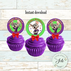 May include: Three purple cupcakes with red bows and round edible toppers featuring a cartoon character with the text "Happy Birthday" in red and white stars. The character is wearing a purple shirt with a letter C on it. The toppers are on a light wood background.