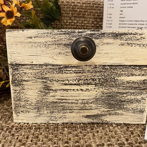 May include: A white wooden box with a black distressed finish and a black metal knob on the lid. The box is sitting on a brown woven surface.