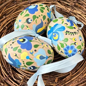 May include: Three hand-painted Easter eggs with blue floral designs and light blue ribbons, nestled in a woven nest. The eggs feature a beige base color with blue, yellow, and green floral patterns, perfect for Easter decorations.