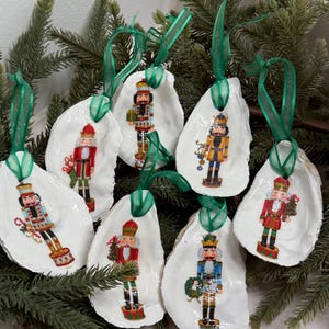 May include: Handmade Christmas decorations featuring nutcracker designs. Each ornament is crafted on a white oyster shell and adorned with a detailed nutcracker figure. Green ribbon loops are attached for hanging, set against a backdrop of evergreen branches.