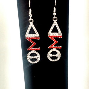May include: A pair of silver-toned dangle earrings. Each earring features a triangle, a sigma, and an omega symbol, all encrusted with clear and red rhinestones. The earrings hang from a simple hook.