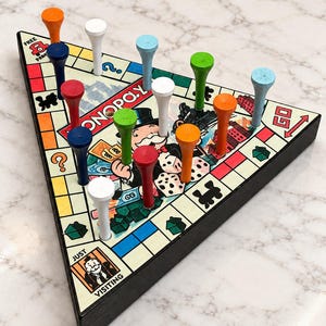 May include: A triangular Monopoly-themed peg solitaire board game featuring a colorful game board with the Monopoly logo, Rich Uncle Pennybags, and various game spaces. The board is populated with multiple wooden pegs in orange, white, blue, red, green, and light blue colors.