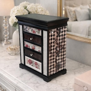 May include: Black jewelry box with five drawers. The drawers have floral designs and crystal knobs. The sides feature a checkered pattern and mirrored accents. The box sits on a marble surface.