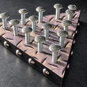 May include: A triangular base with a dark, diamond-plate metal top surface featuring fifteen vertical silver carriage bolts arranged in a grid. The sides of the base are black with silver rivets spaced evenly along the edges.