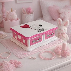 May include: A white and pink jewelry box with two drawers, adorned with a Snoopy illustration holding a heart. The box is surrounded by pink accessories, including bracelets, earrings, and nail polish, creating a charming, feminine aesthetic.