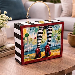 May include: A rectangular handbag with black and white stripes on the sides and a gold chain handle. The front features a colorful painting of legs in striped stockings and red high heels. The bag has a red border.