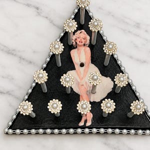 May include: A triangular black wall hook rack featuring a central image of Marilyn Monroe in a white dress. The rack is decorated with a border of small white beads and twelve silver-toned hooks, each topped with a circular faux pearl and rhinestone flower accent.