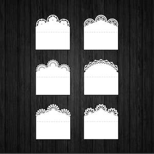 May include: Six white, decorative paper cutouts with intricate lace-like edges. Each cutout has a rectangular base with a scalloped or arched top. The cutouts are arranged on a dark wood-grain background, suitable for crafting or scrapbooking.