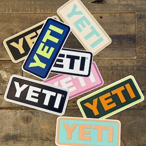 May include: A collection of rectangular YETI stickers in various color combinations, including black and gold, blue and yellow, and pink and white. The stickers are arranged on a wooden surface.