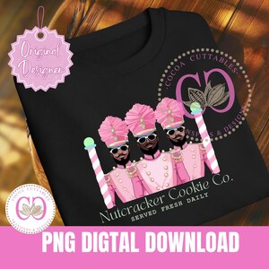 Pode incluir: Três soldados quebra-nozes com uniformes rosa, chapéus pretos e acessórios de bengala de doces. O texto "Nutcracker Cookie Co. Served Fresh Daily" está abaixo dos soldados.
