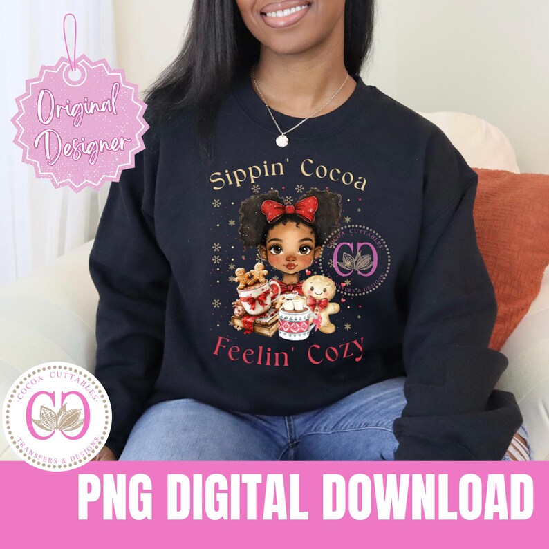 K&ouml;nnte beinhalten: Ein schwarzer Pullover mit dem Text "Sippin' Cocoa Feelin' Cozy" in Pink. Eine Cartoon-Illustration eines schwarzen M&auml;dchens mit braunen Haaren, das eine rote Schleife tr&auml;gt, eine Tasse hei&szlig;e Schokolade h&auml;lt und von weihnachtlichen Dekorationen umgeben ist.