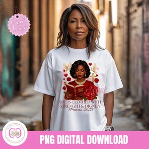May include: White t-shirt featuring a graphic of a woman reading a book, with doves, roses, and a cross. The text reads "SHE IS CLOTHED WITH STRENGTH & DIGNITY Proverbs 31:25". The shirt has a round neckline and short sleeves.
