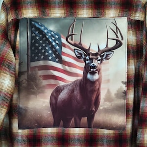 May include: A red, white, and blue American flag is behind a brown and white deer with large antlers. The image is on a brown, white, and red plaid fabric.