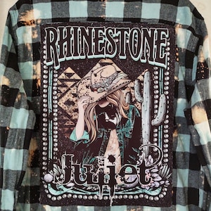 May include: A blue and black plaid flannel shirt with a graphic of a woman wearing a cowboy hat and the text "Rhinestone Juliet" on the back.
