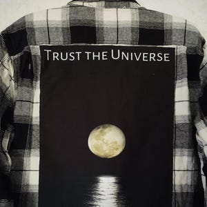 May include: Black and white plaid flannel shirt with a black patch on the back. The patch features a full moon over a body of water and the text "TRUST THE UNIVERSE".