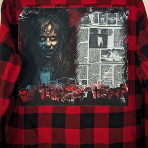 May include: A red and black buffalo plaid flannel shirt featuring a large rectangular patch on the back. The patch displays a graphic of a character with glowing yellow eyes, a newspaper clipping titled The Exorcist, and a graveyard silhouette.