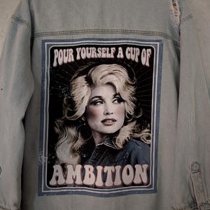 May include: A light-wash denim jacket featuring a large rectangular graphic on the back. The print displays a portrait of a blonde person with the text "POUR YOURSELF A CUP OF AMBITION" in a retro font. The jacket has distressed details and rhinestone cuffs.