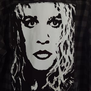 May include: A black and gray plaid button-down shirt with a large white and black graphic of a woman's face on the back. The graphic is a stylized portrait with bold lines and a striking contrast.