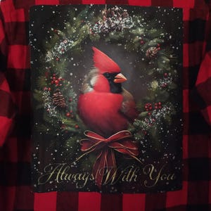 May include: Red and black plaid flannel shirt with a graphic of a cardinal bird in a wreath. The cardinal is red with a black mask and is surrounded by a wreath of pine needles, berries, and pine cones. The text "Always With You" is below the bird.
