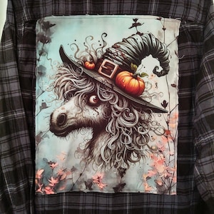 May include: A black and gray plaid flannel shirt with a patch sewn on the back. The patch features a white, furry creature with a witch's hat and pumpkins. The background of the patch is a light blue watercolor with pink flowers.