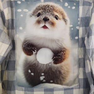 May include: A light blue and white plaid button-up shirt with a patch featuring a cute otter holding a snowball in a snowy scene. The otter has brown fur and is surrounded by falling snow.