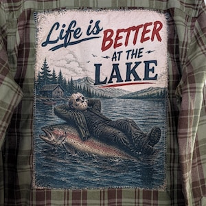 May include: A green and brown plaid flannel shirt featuring a large rectangular patch on the back. The patch displays a graphic of a figure in a hockey mask reclining on a giant fish in a lake, with the text "Life is BETTER AT THE LAKE".
