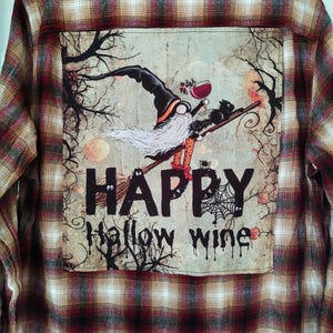 May include: A red and black plaid flannel shirt with a patch on the back. The patch features a cartoon gnome with a black hat and white beard riding a broomstick with a black cat. The text on the patch reads "HAPPY HALLOW WINE".