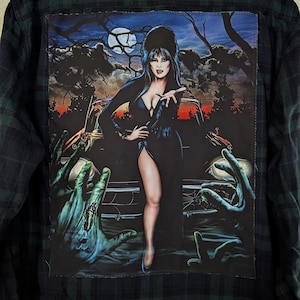 May include: A dark green and black plaid flannel shirt featuring a large rectangular patch on the back. The patch displays a graphic of a woman with long black hair standing in front of a car at night, surrounded by reaching zombie-like hands.