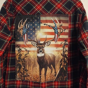 May include: A red and black plaid flannel shirt with a patch on the back. The patch features a deer standing in a field with an American flag in the background.