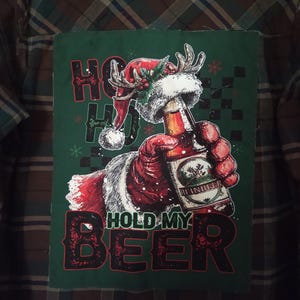 May include: Plaid button-down shirt with a green patch featuring Santa Claus holding a beer bottle. The patch reads "HO HO HOLD MY BEER" in red and white text. The shirt is a mix of brown, green, and black.