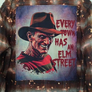 May include: A brown and tan plaid flannel shirt with a graphic of Freddy Krueger. The graphic features Freddy's face, a brown hat, and the text "EVERY TOWN HAS AN ELM STREET" in red and white.