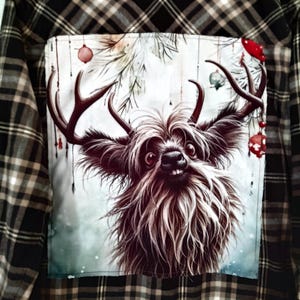 May include: A black and brown plaid flannel shirt with a festive patch on the back. The patch features a furry, cartoon-like creature with antlers, surrounded by pine branches and red ornaments.