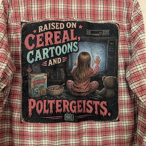 May include: A pink and white plaid button-down shirt featuring a large graphic on the back. The graphic depicts a child sitting before a static-filled television screen with the text "RAISED ON CEREAL, CARTOONS AND POLTERGEISTS" and a cereal box.