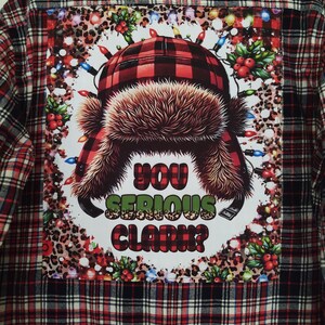 May include: Red and black plaid flannel shirt with a festive patch on the back. The patch features a cartoon image of a person wearing a red and black plaid hat with a fur trim, surrounded by Christmas lights and holly. The text "You Serious Clark?" is printed on the patch.