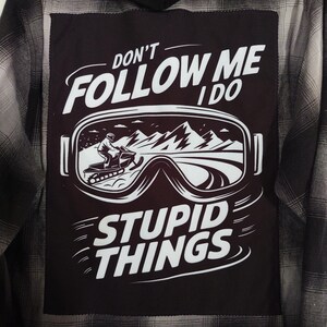 May include: A hooded flannel shirt in shades of gray and black. The back features a black patch with white text that reads "DON'T FOLLOW ME I DO STUPID THINGS." The patch also includes a graphic of a person on a snowmobile.