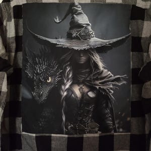 May include: Black and white plaid flannel shirt with a large graphic on the back. The graphic depicts a witch with a dragon, both in shades of gray. The witch wears a pointed hat and dark clothing.