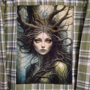 May include: A green and white plaid flannel shirt with a large image of a woman with long brown hair and a tree growing out of her head. The image is surrounded by a dark green border.