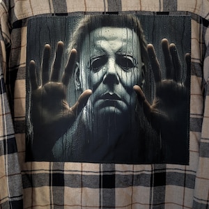 May include: A long-sleeve button-down shirt featuring a tan, black, and white plaid pattern. The back of the shirt displays a large, square graphic print of a pale, masked figure with dark eyes, appearing behind a wet, rain-streaked glass surface.